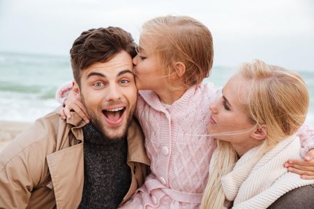 Three people smiling and hugging outdoors at the beach, one child kissing a man's cheek