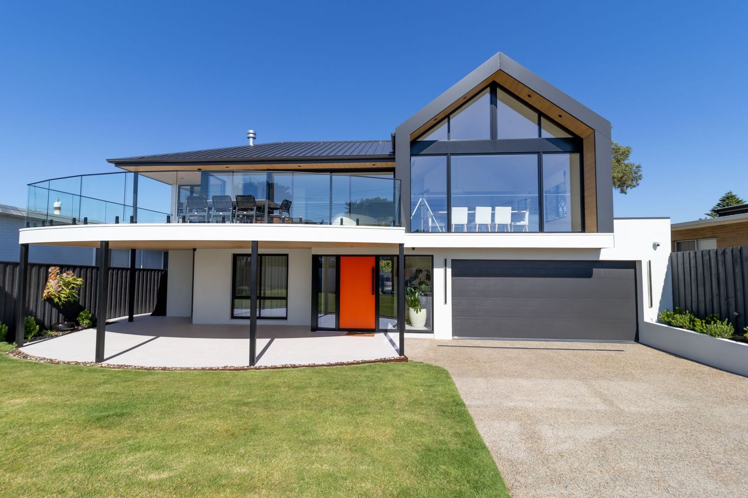 Modern two-story house with a white exterior, large glass balcony, gable roof feature, and a bright orange front door.