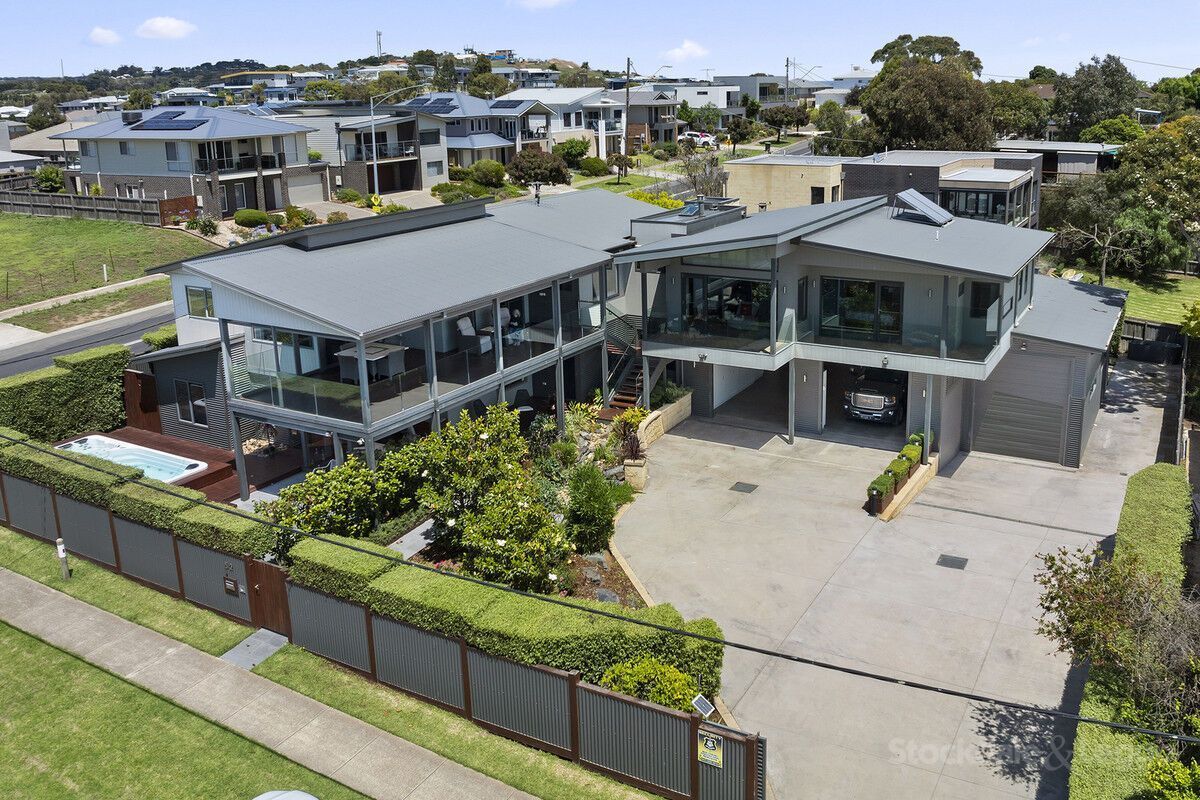 An elevated two-story modern house with grey siding, a balcony, a swimming pool, and a paved driveway.
