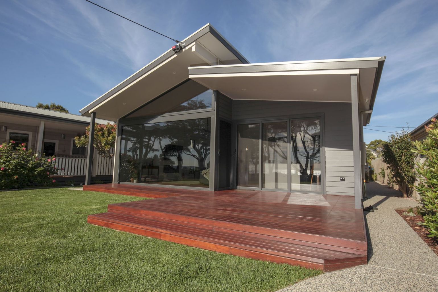 Modern house with a slanted roof, dark gray siding, and a wooden deck overlooking a green lawn under a sunny blue sky.