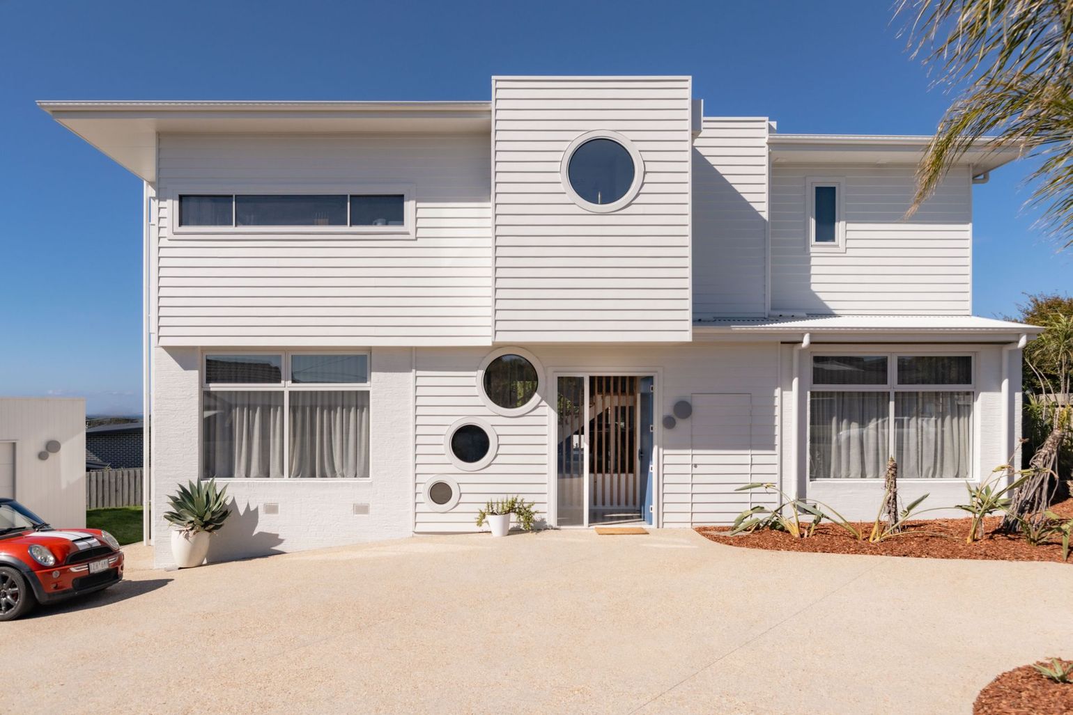 A modern, two-story white house with horizontal siding, round porthole windows, and a paved driveway.