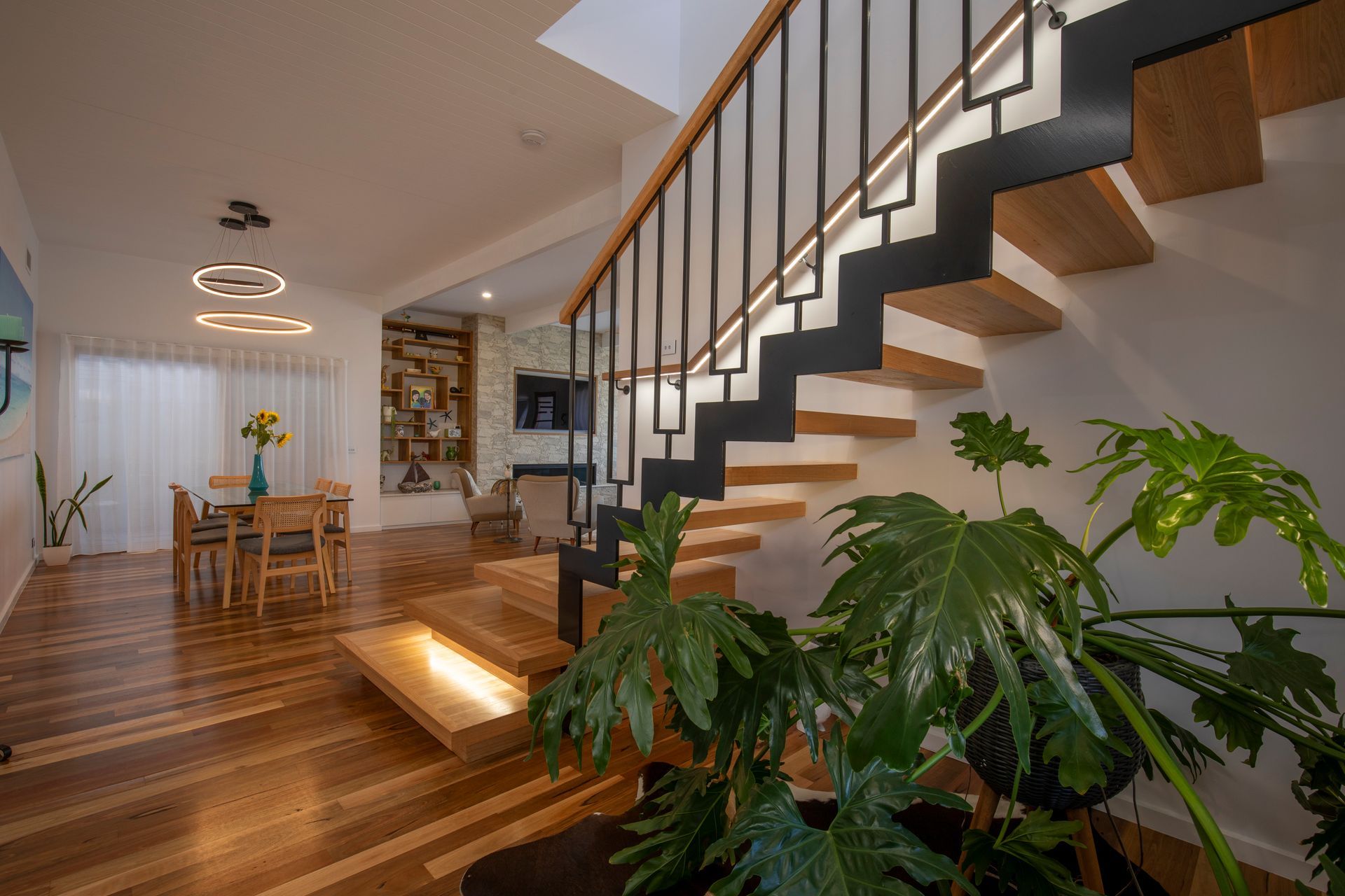 Modern staircase with light wood steps and black railings overlooking a dining area with a table, chairs, and plant.