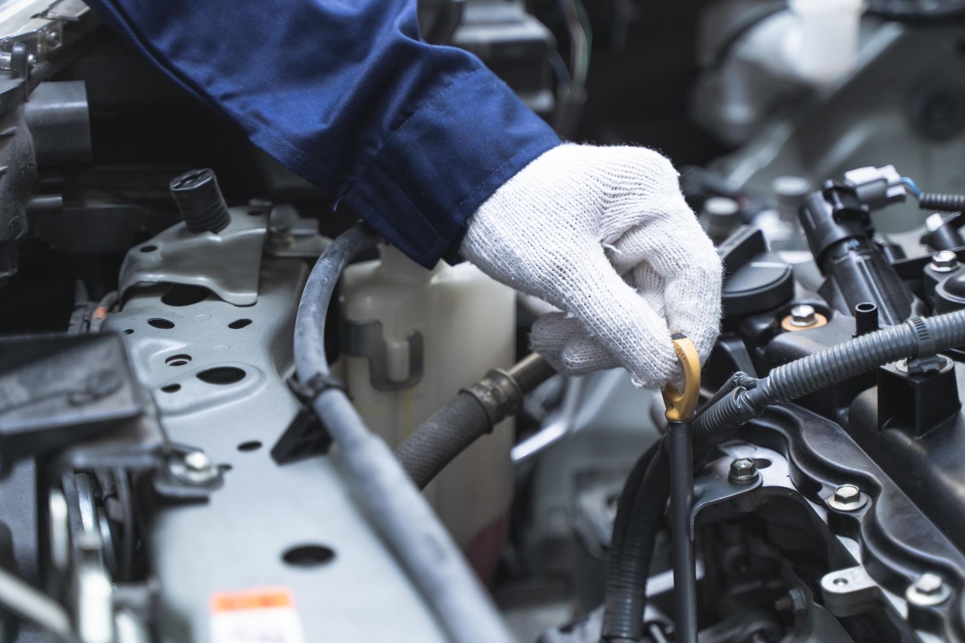 Mechanic's gloved hand checking oil level in a car engine bay. Blue sleeve, white glove, gray engine.