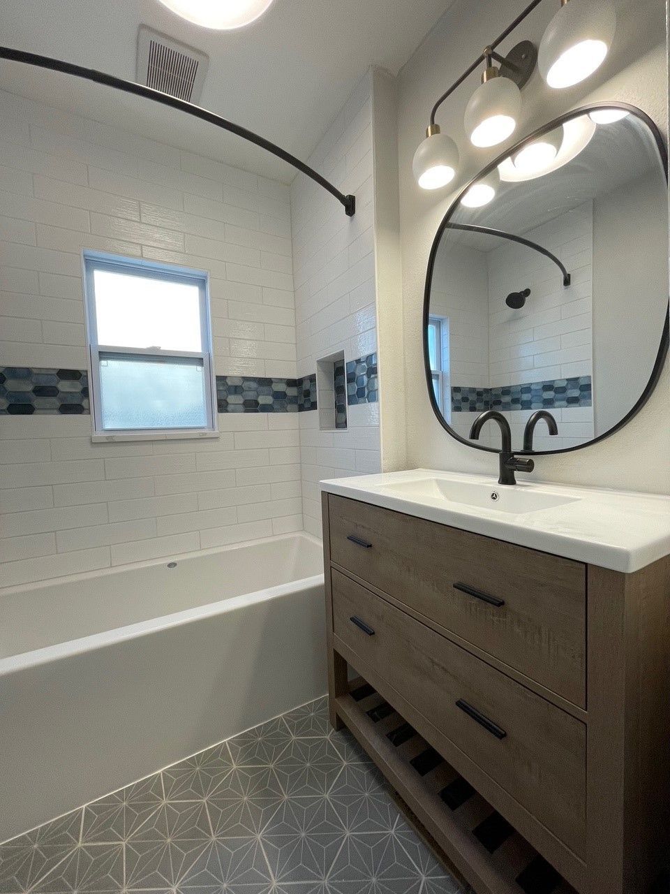 Bathroom with a wooden vanity, oval mirror, and white and blue tiled walls. A window and a bathtub are also visible.