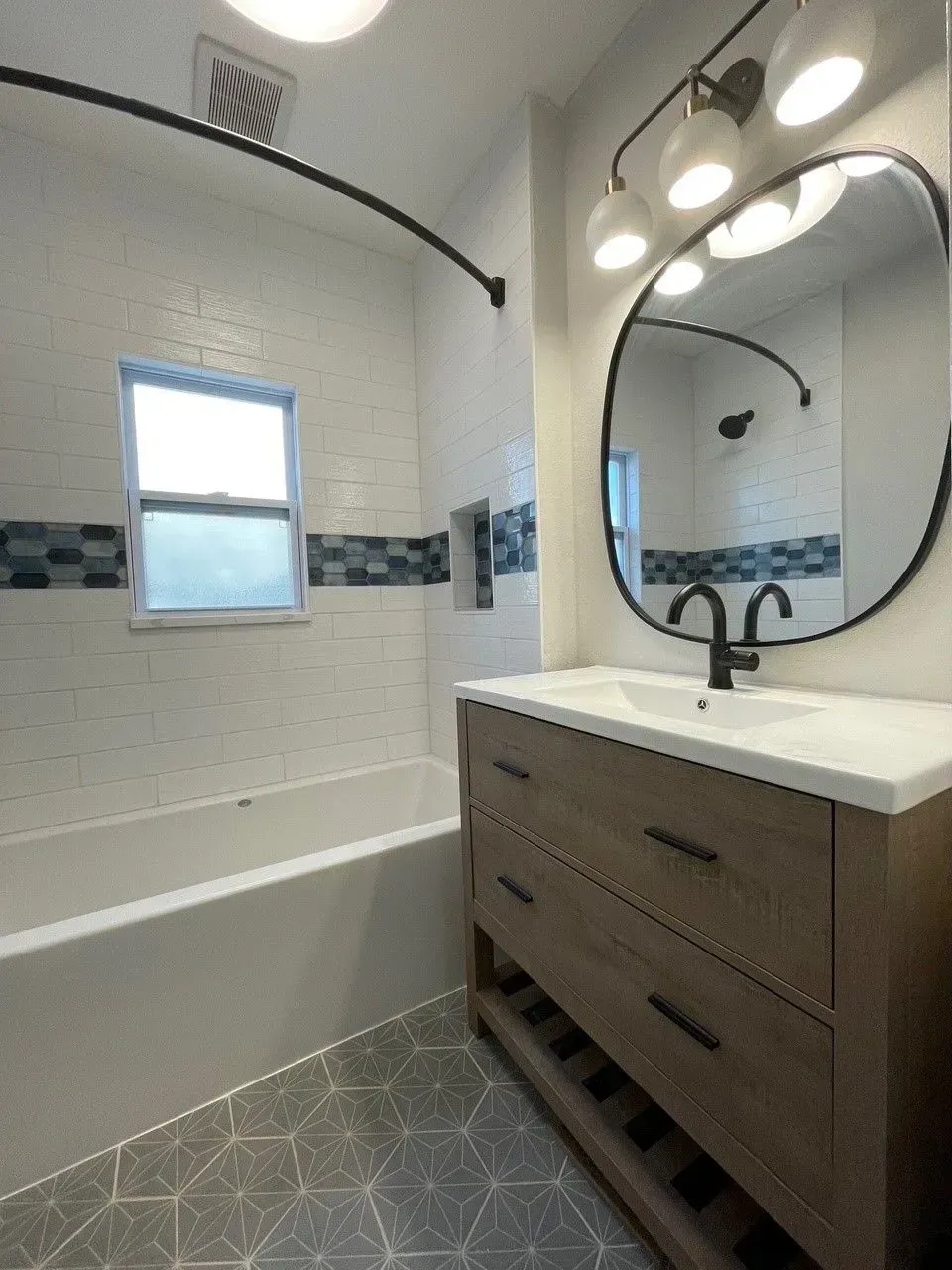 Modern bathroom with a white tub, wooden vanity, oval mirror, and geometric floor tiles.