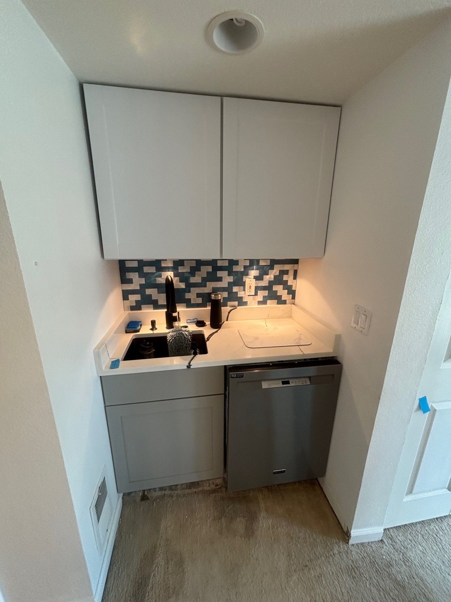 A small, modern kitchen area with white cabinets, gray countertop, and a black sink and faucet. A dishwasher is to the right.