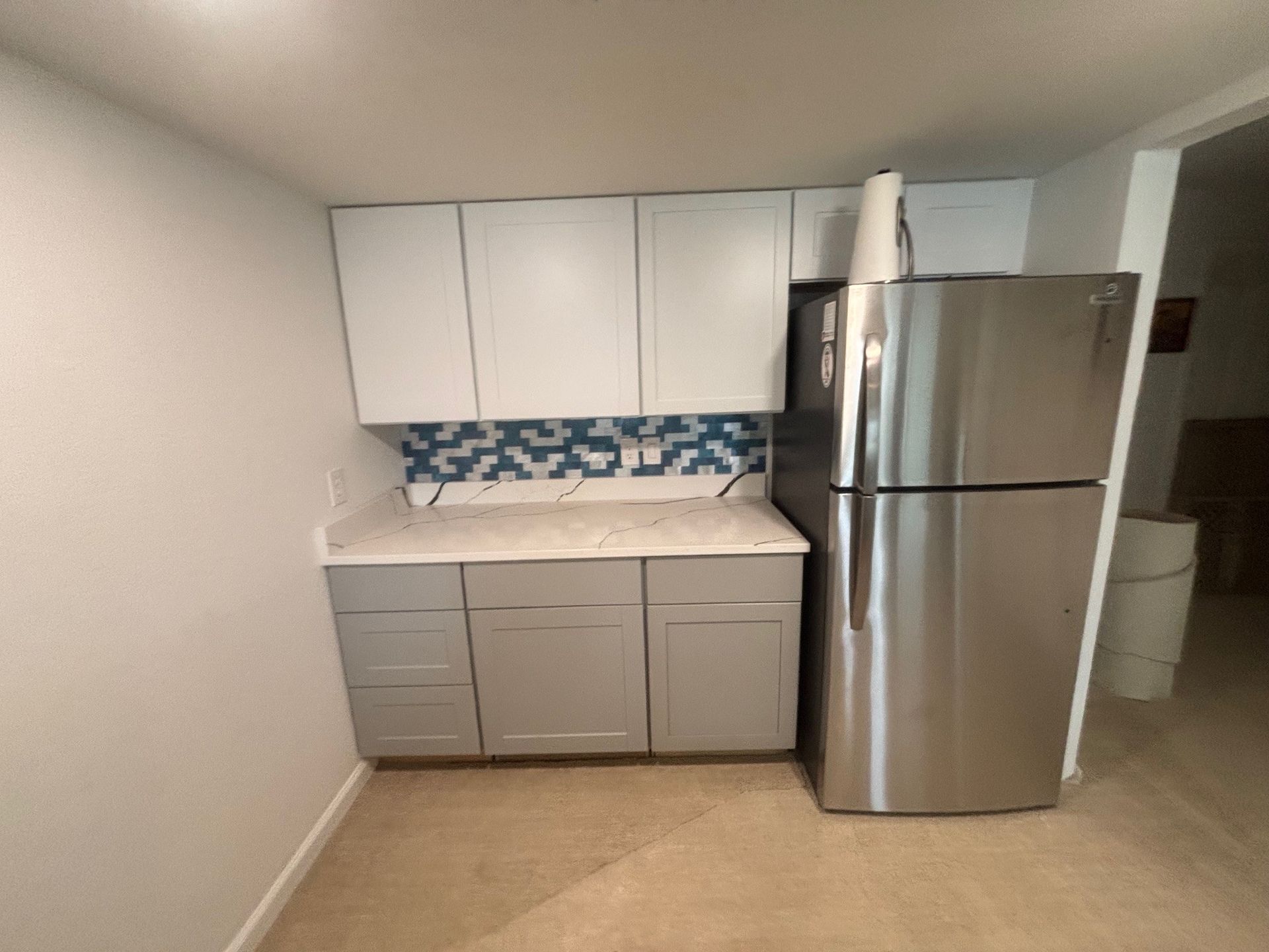 kitchenette with light gray cabinets, white countertops, and a blue and white tile backsplash. A stainless steel refrigerator