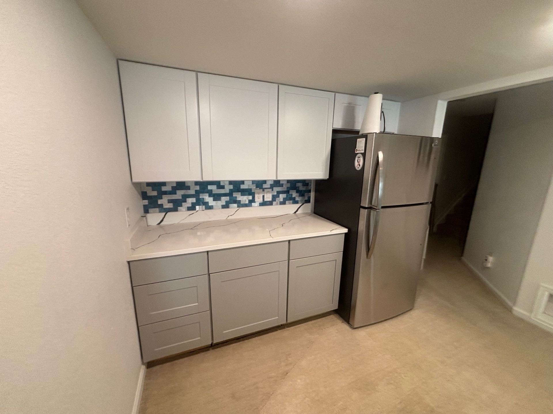 A small kitchen with gray cabinets, a stainless steel refrigerator, and a blue and white tiled backsplash.