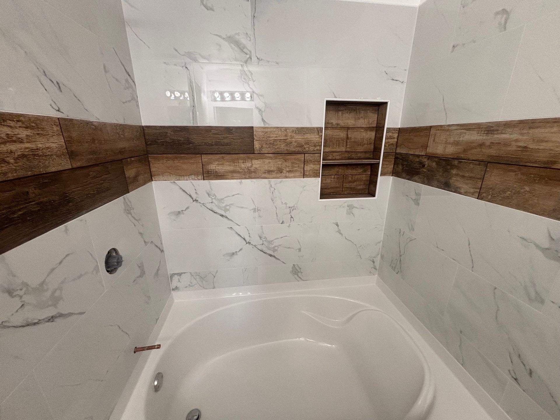 Bathroom with marble-look white tiles and wood-look accent tiles. A white oval bathtub sits below a built-in shelf.