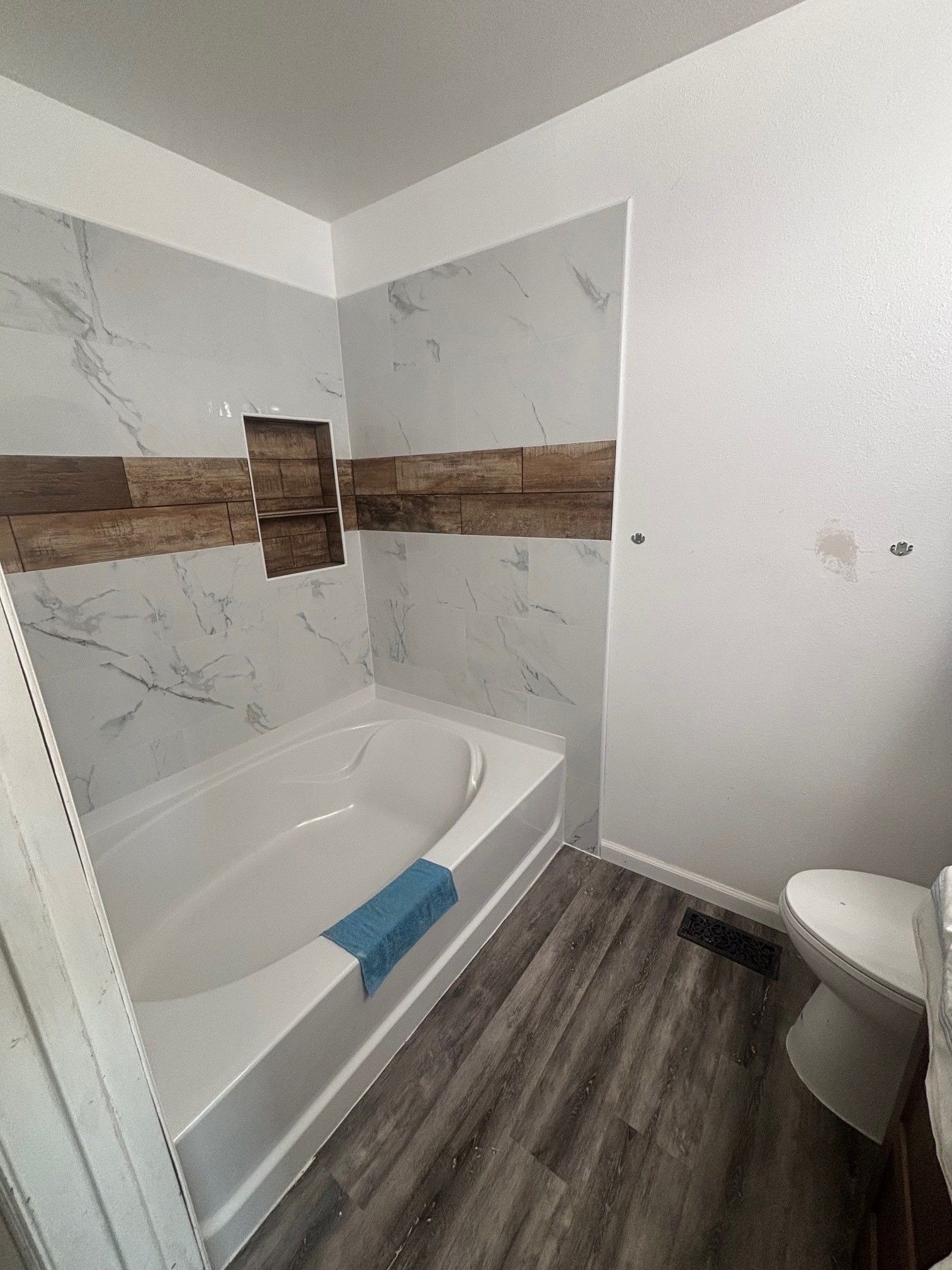 Bathroom with a white tub, marble tile, wooden accent, and gray wood-look flooring. A toilet is in the corner.