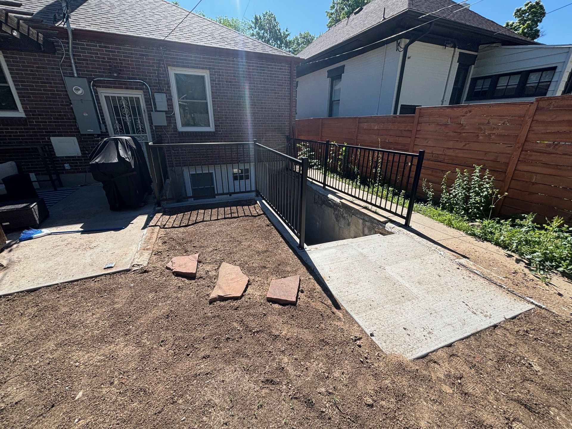 A paved ramp with black railings leads down to a basement door. The ramp is in a backyard with brown mulch.