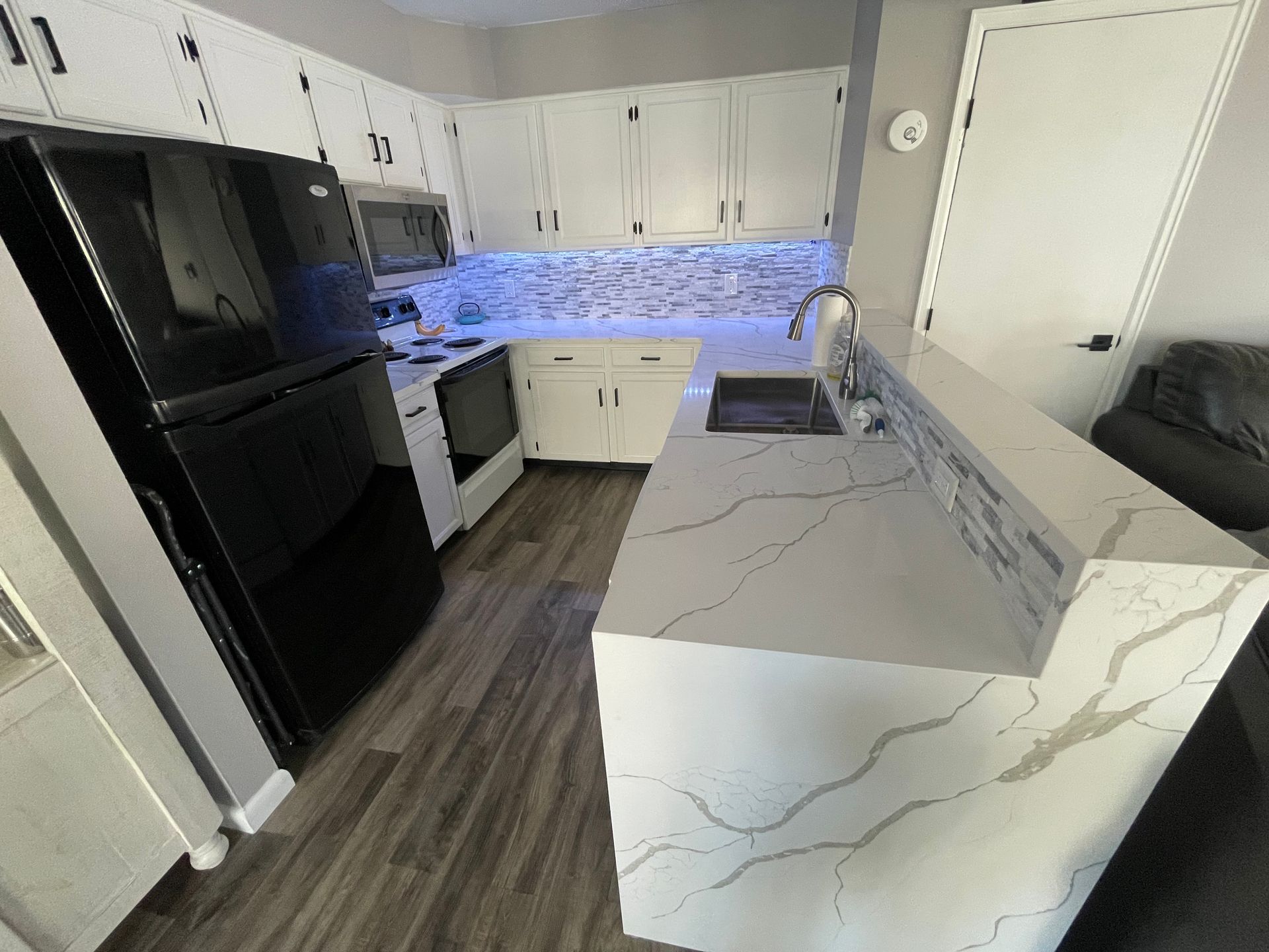 A kitchen with white cabinets, light-colored countertops with grey veining. Wooden flooring and a backsplash