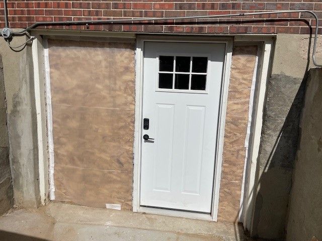 White door in a brick and concrete basement entry. Plywood surround with electrical wiring.