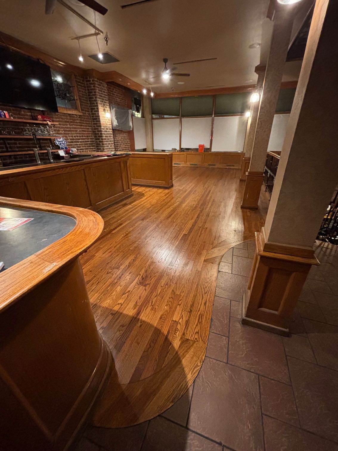 Empty bar interior with a wooden floor, bar, and brick wall. Natural light streams in, illuminating the space.