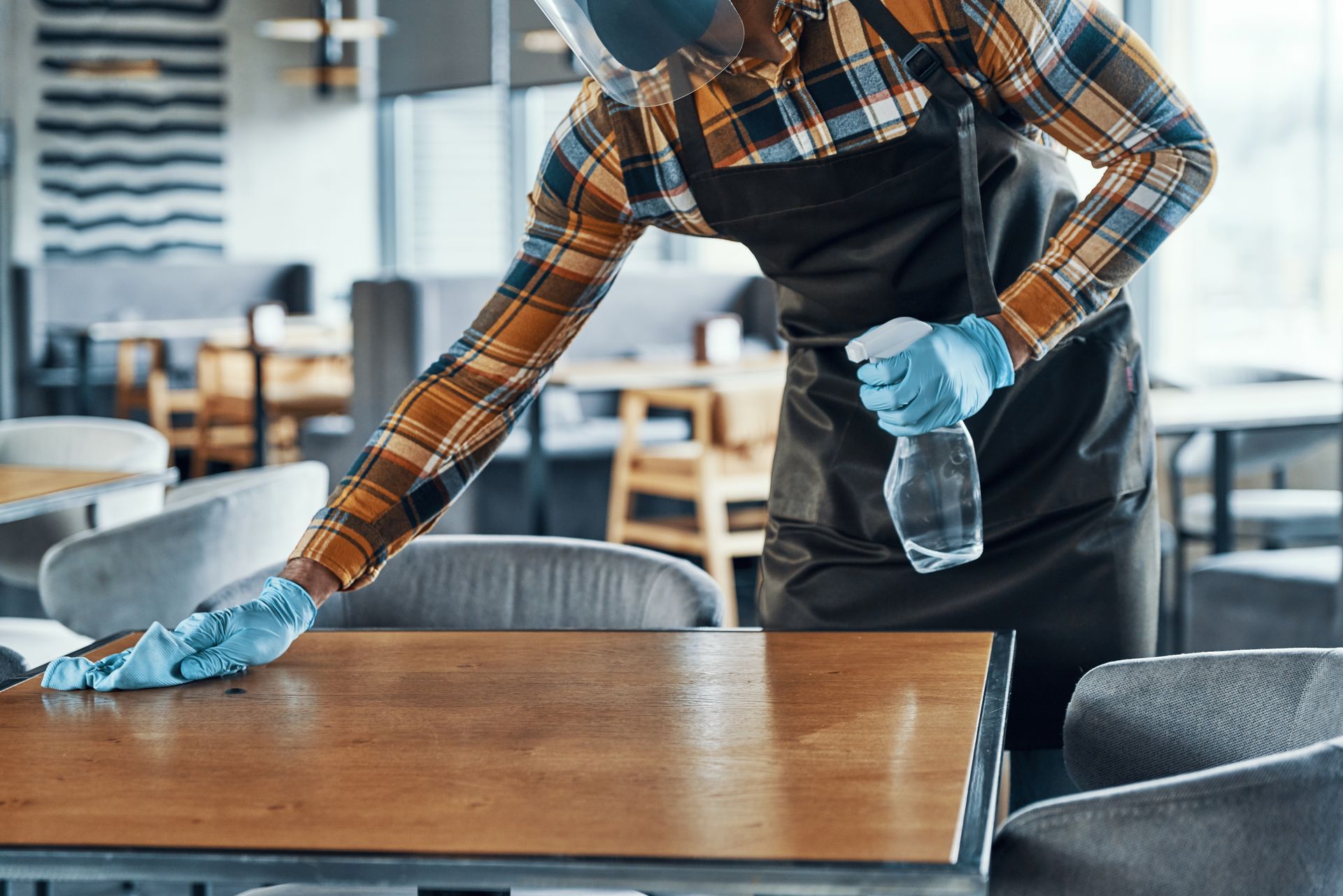 A man wearing a mask and gloves is cleaning a table in a restaurant.