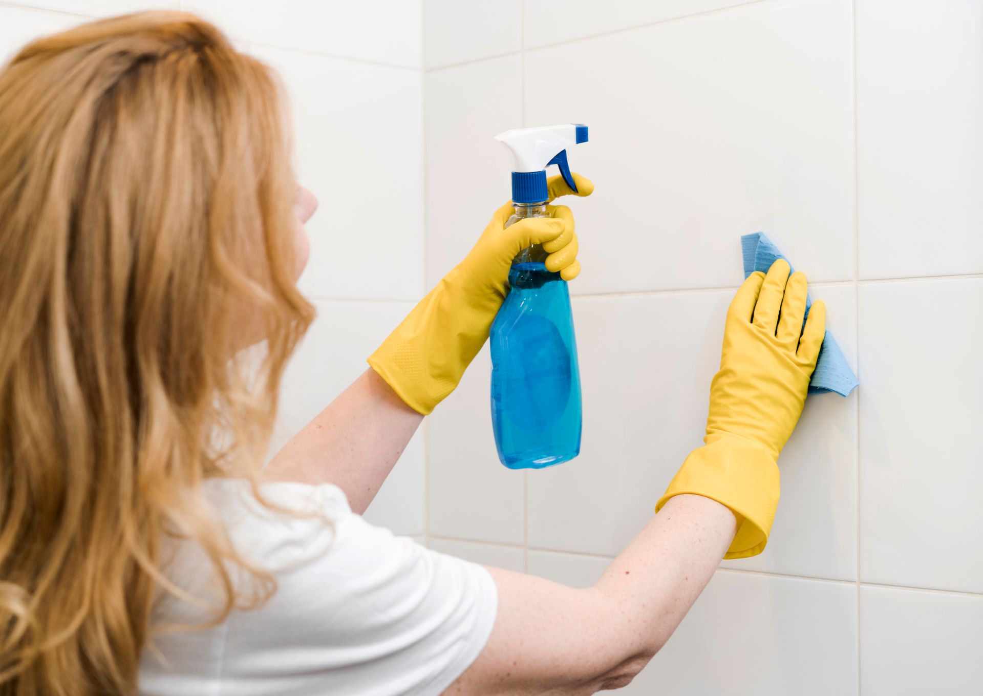 A woman wearing yellow gloves is cleaning a wall with a spray bottle and a cloth.