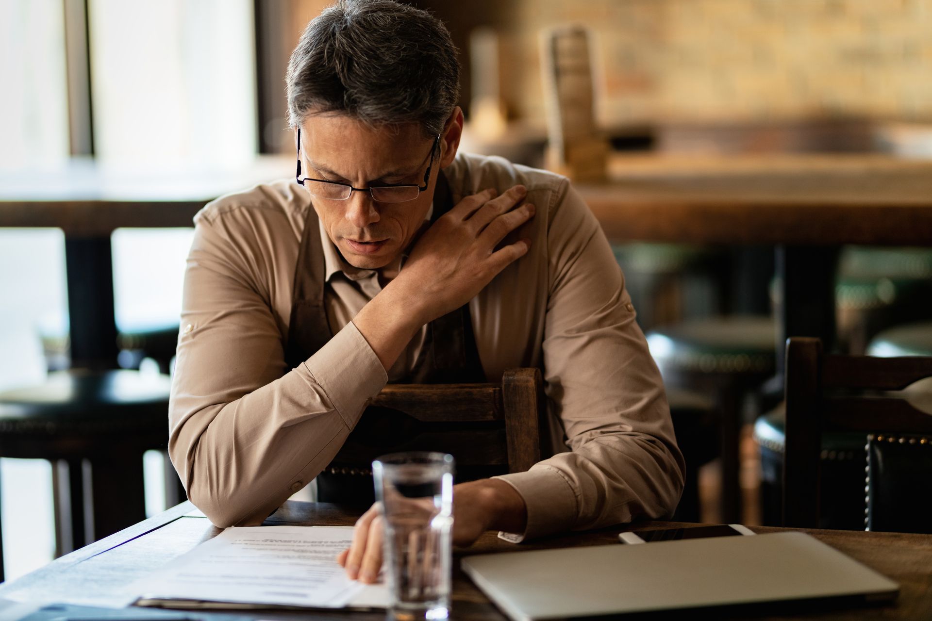 A person in a tan shirt sits at a table, looking down at documents with a hand to their chin in a dimly lit restaurant.