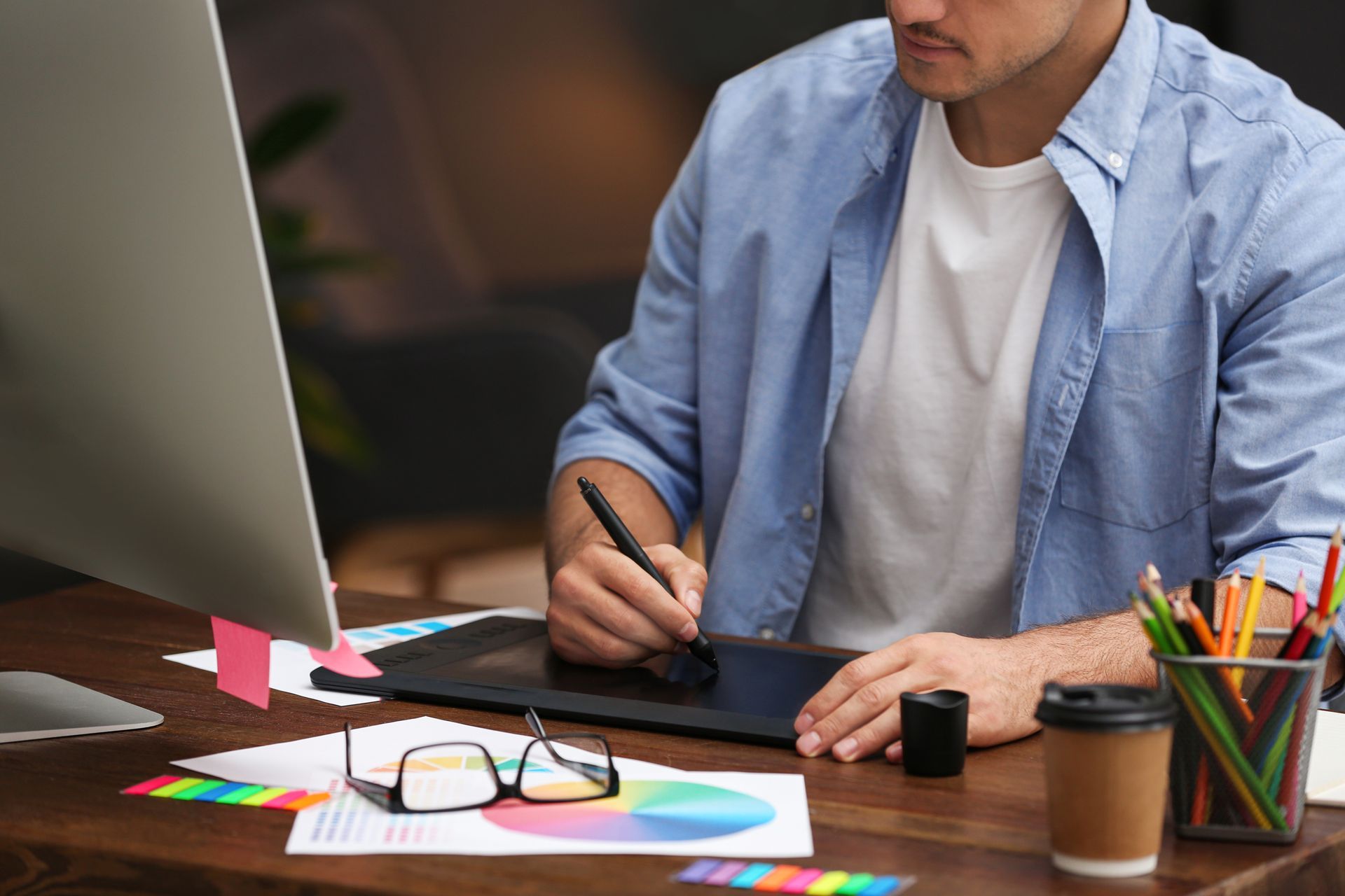 A person in a light blue shirt uses a digital drawing tablet at a desk with a computer monitor, glasses, and pens.