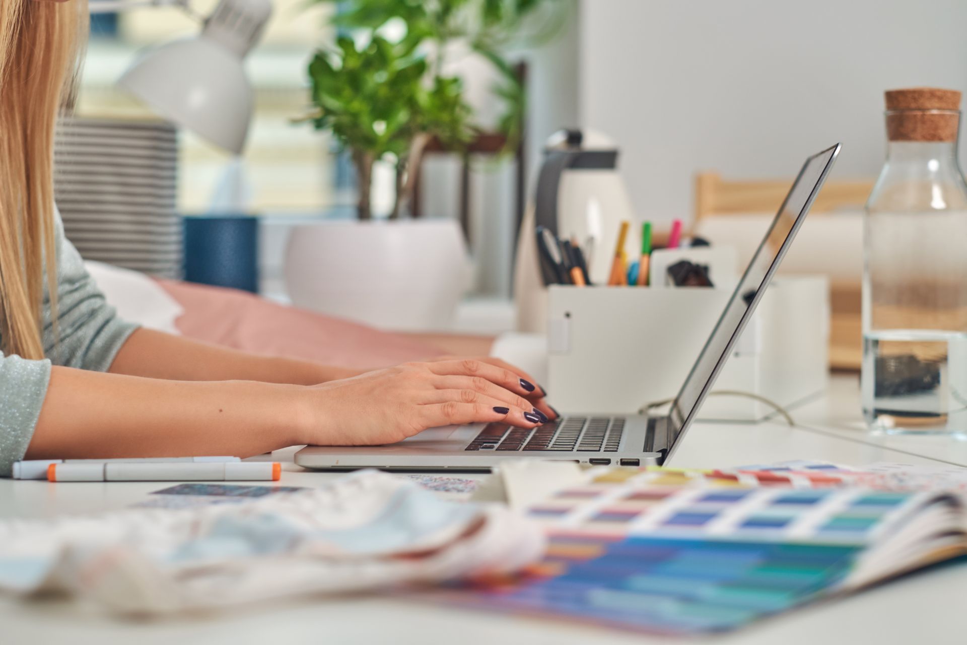 A person types on a laptop at a workspace with a color swatch book, a desk lamp, a plant, and a glass water bottle.