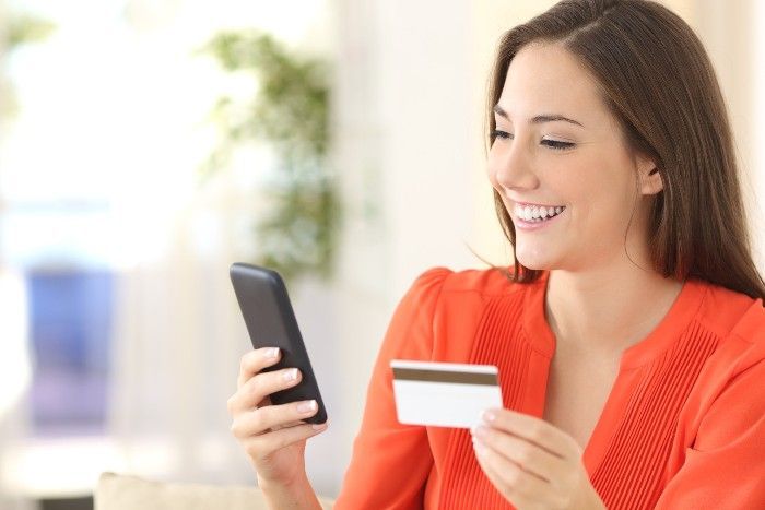 Woman smiling, holding a phone and credit card, possibly shopping online. Orange shirt, indoor setting.