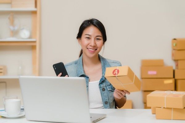 箱に囲まれたノートパソコンのそばで、荷物と携帯電話を持ちながら微笑む女性。