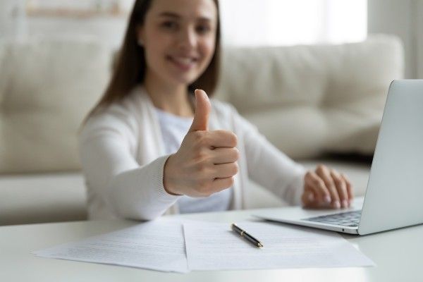 ノートパソコンと書類を持ってテーブルに座り、親指を立てている女性。