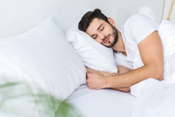 Man asleep in white bed, wearing a white t-shirt, smiling slightly.