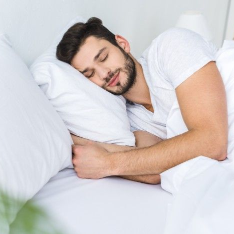 Man asleep in white bed, wearing a white t-shirt, smiling slightly.