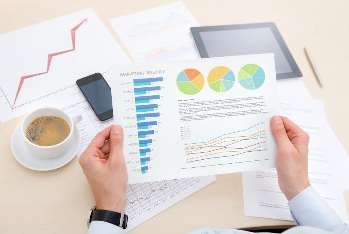 Person holding financial charts, surrounded by papers, tablet, phone, and coffee.