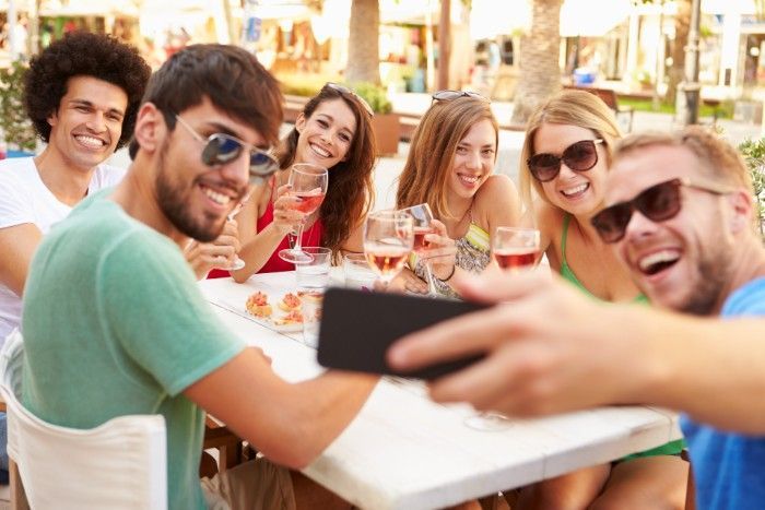 Group of friends smiling and taking a selfie at an outdoor restaurant, holding drinks.