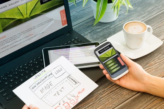 Person using phone, laptop, and tablet, reviewing a website design with coffee on wooden table.