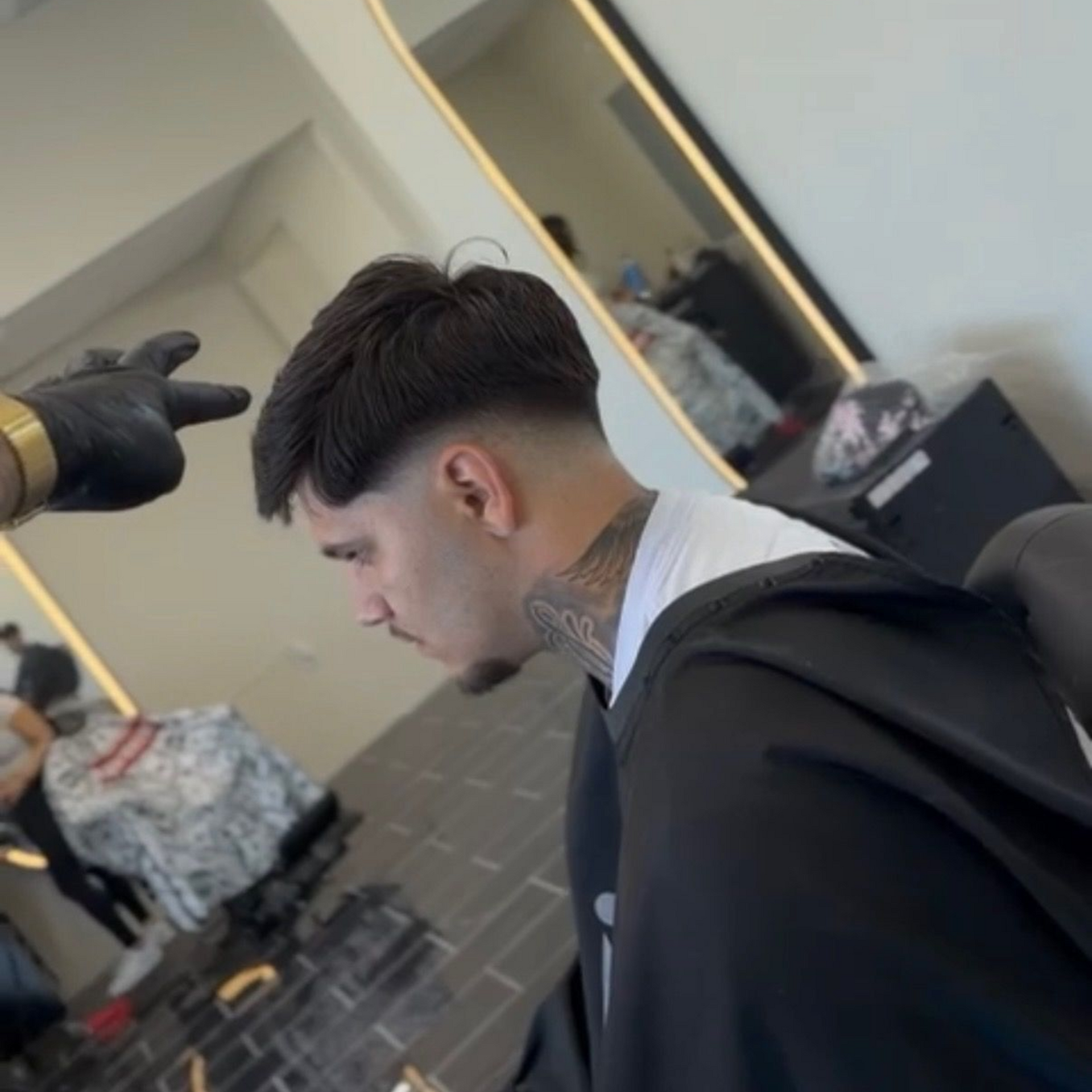 A barber’s gloved hand points toward a client’s mid-fade haircut and neck tattoo in a barbershop setting.