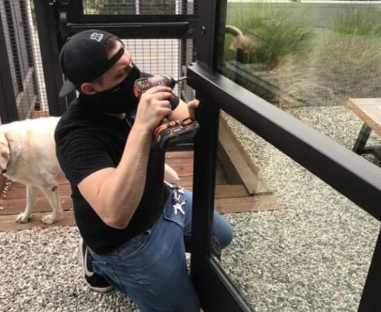 Person in black shirt and mask using a drill on black window frame outdoors. Dog watches.