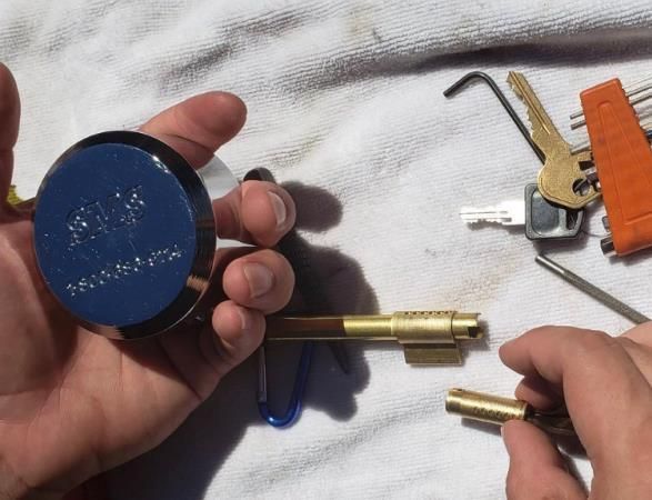 Hands hold a blue padlock and key parts, with other keys and tools on a white cloth.