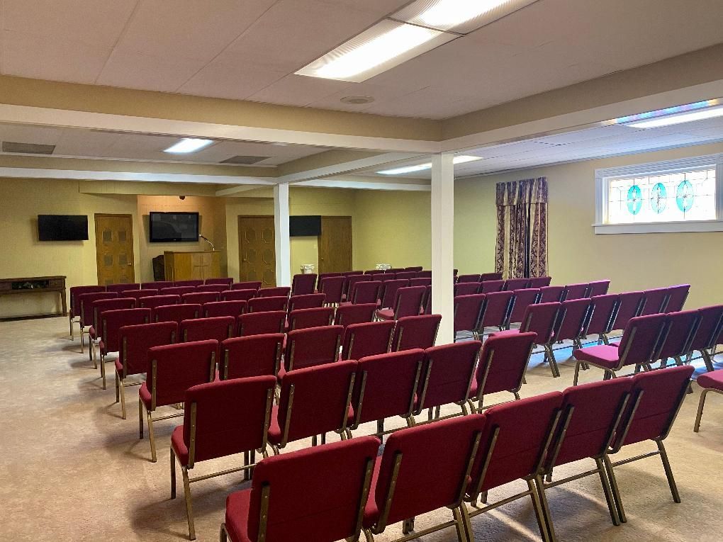 Rows of red chairs in a beige-walled room, likely a meeting space. Three mounted TVs.