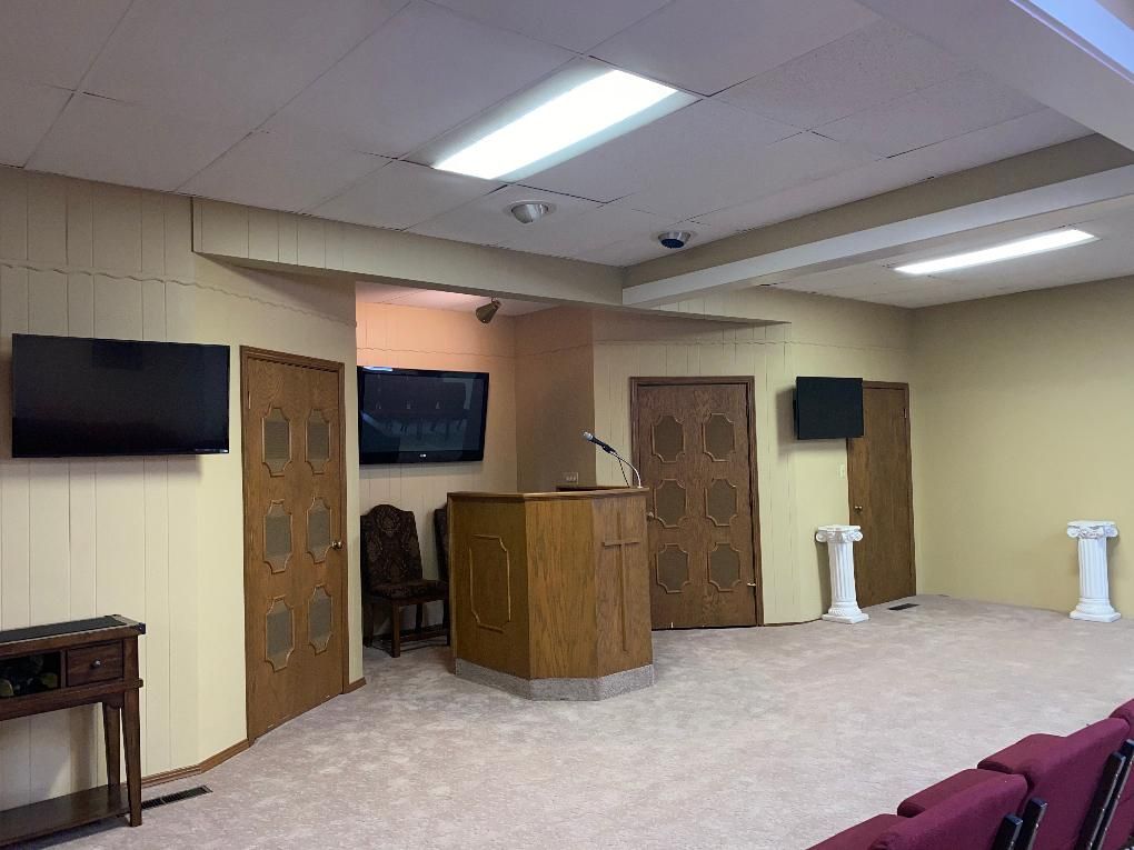 Interior of a room with a wooden podium, doors, and two TVs. Beige walls and carpet.