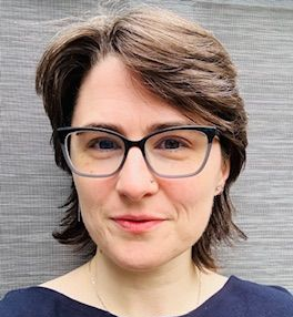 Portrait of a person with short brown hair and black glasses, smiling slightly against a gray background