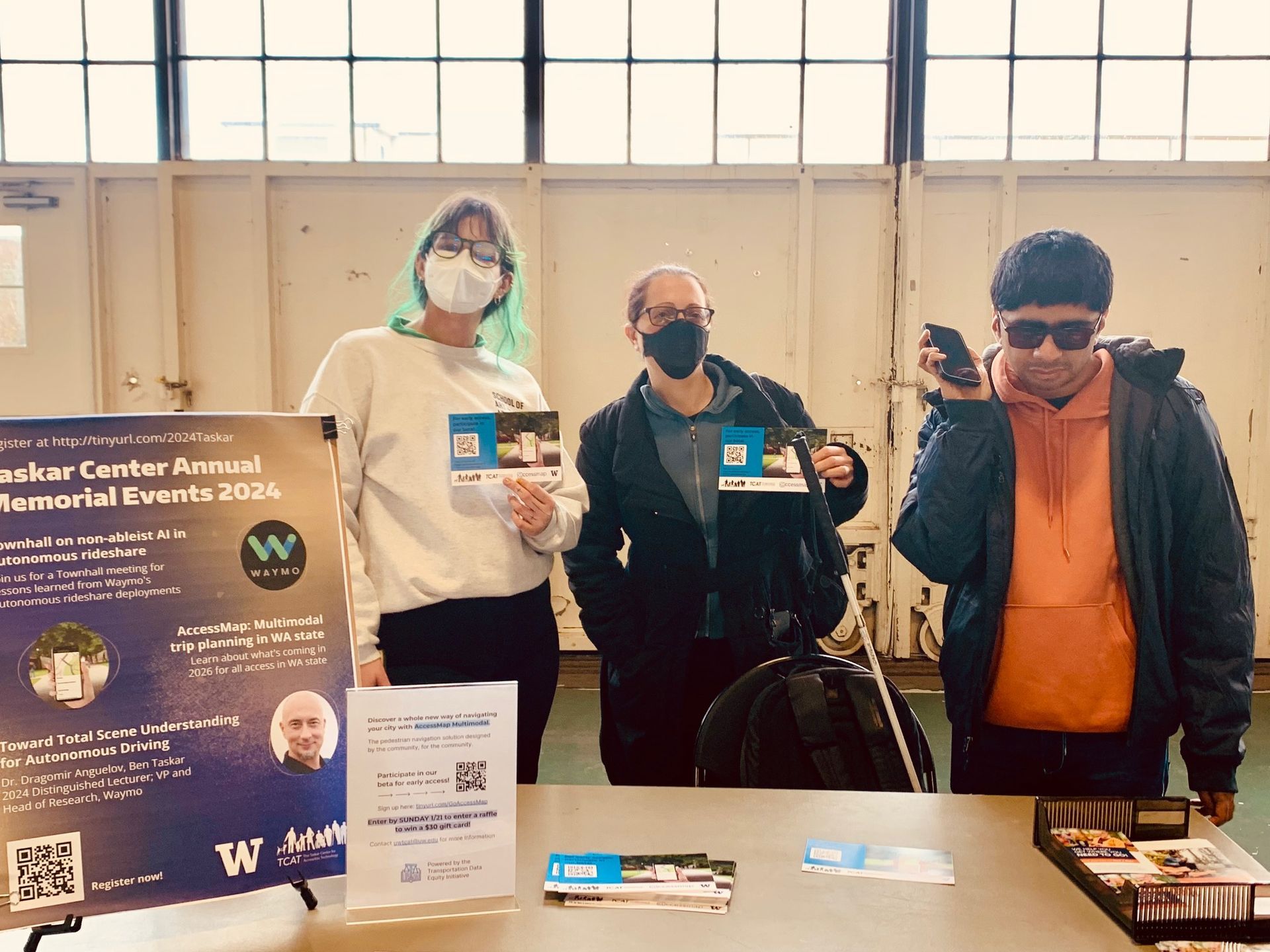 Three people, one with a white cane, in front of a table that has information about AccessMap.