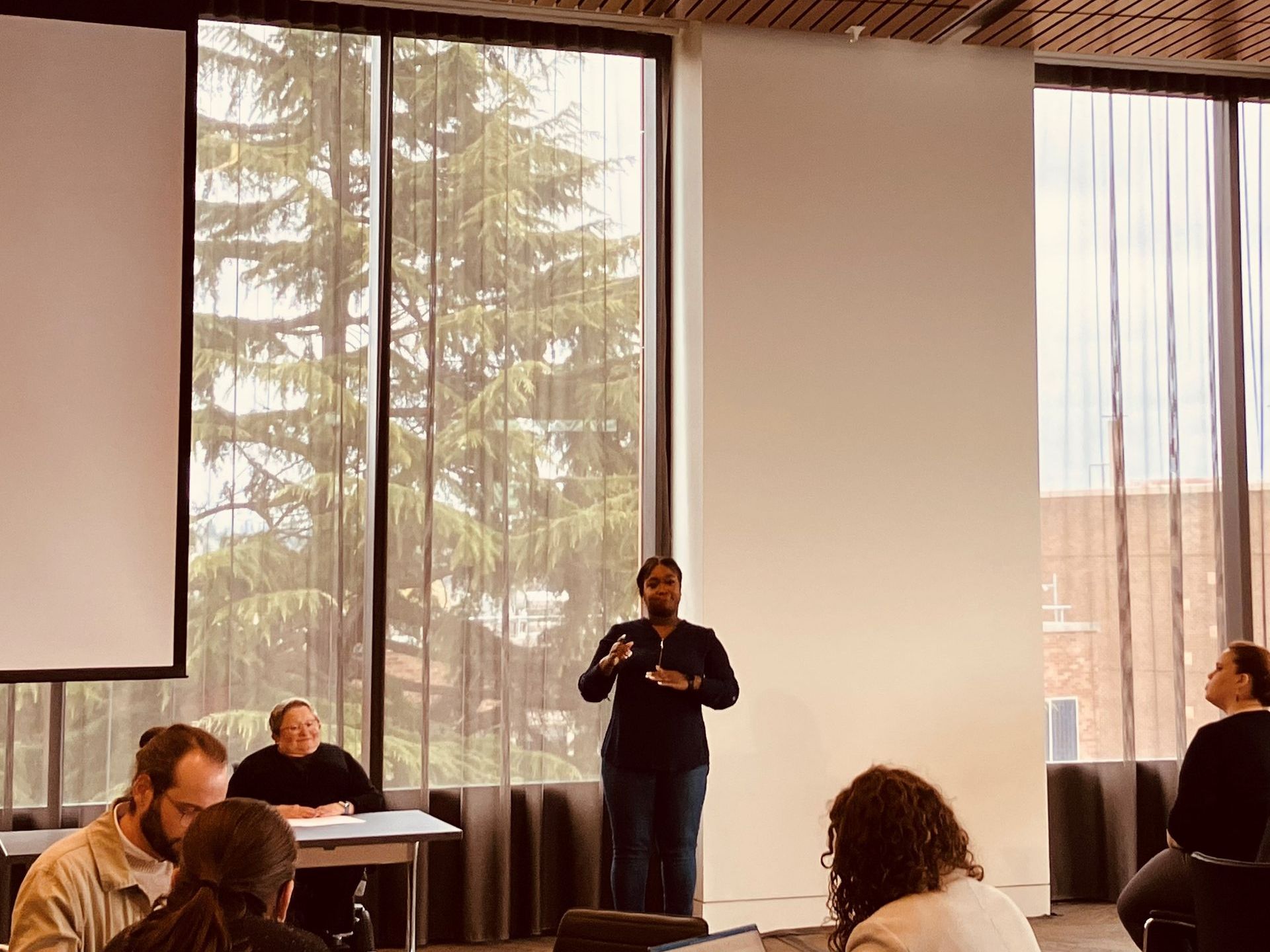 A person wearing black, a sign language interpreter, translates keynote speaker Karen Braitmayer, who is seated at a table in a wheelchair.
