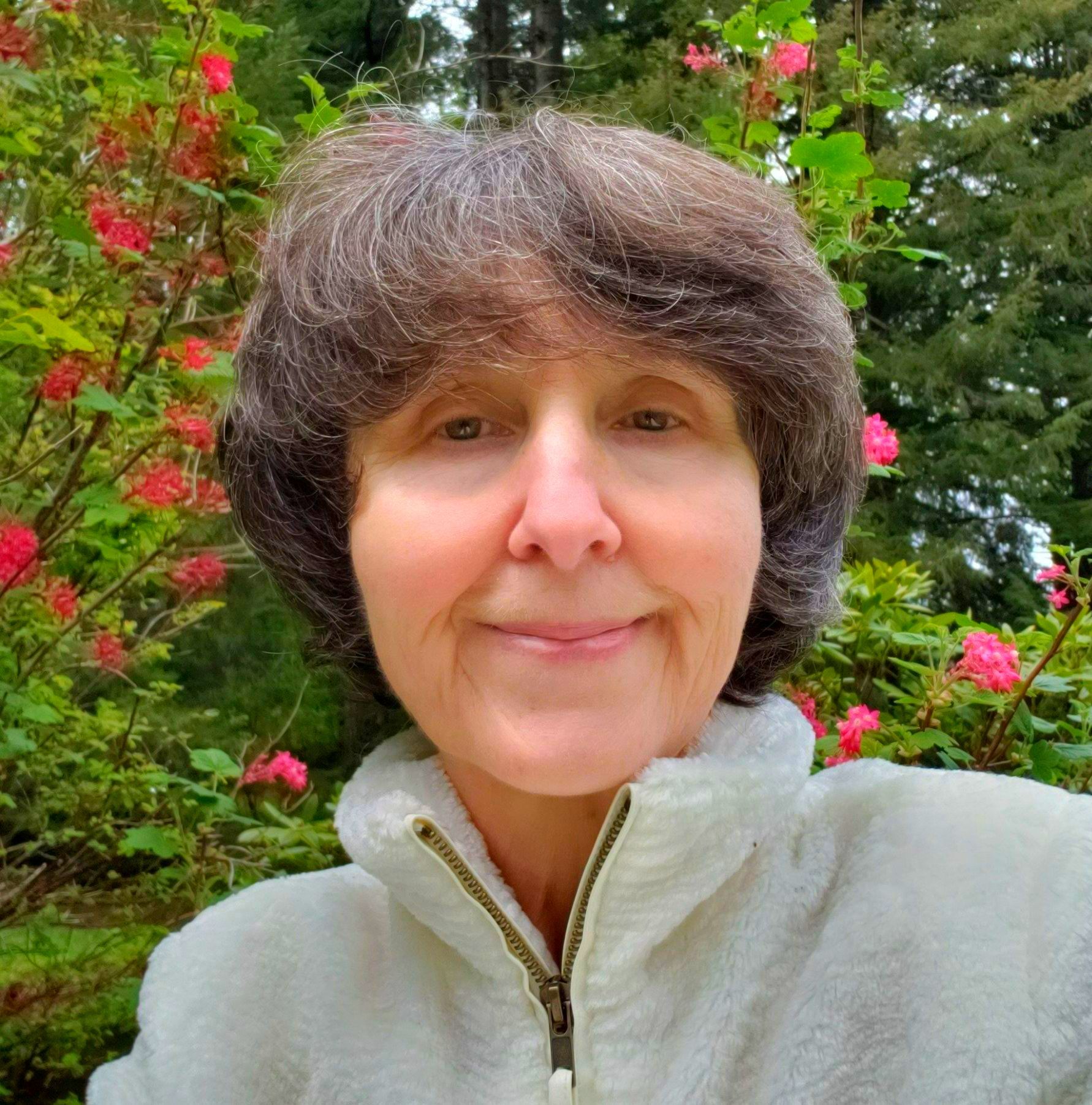Smiling person in a light jacket standing in a garden with pink flowers and green trees