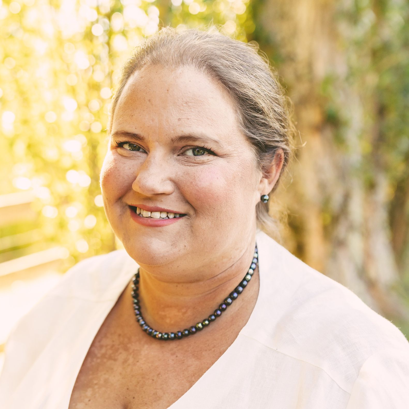 Smiling woman in a white top and beaded necklace, outdoors with a softly blurred golden background