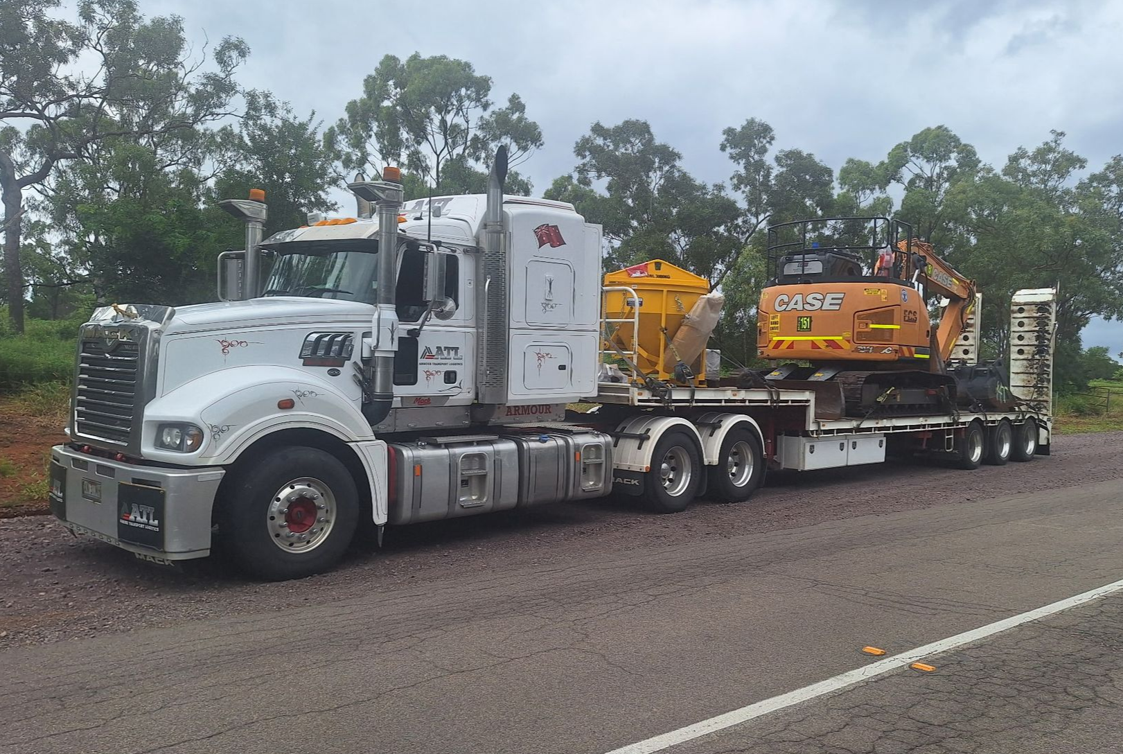 White semi truck hauling yellow heavy equipment on a flatbed trailer beside a road. - Armour Transport Logistics In Rutherford, NSW