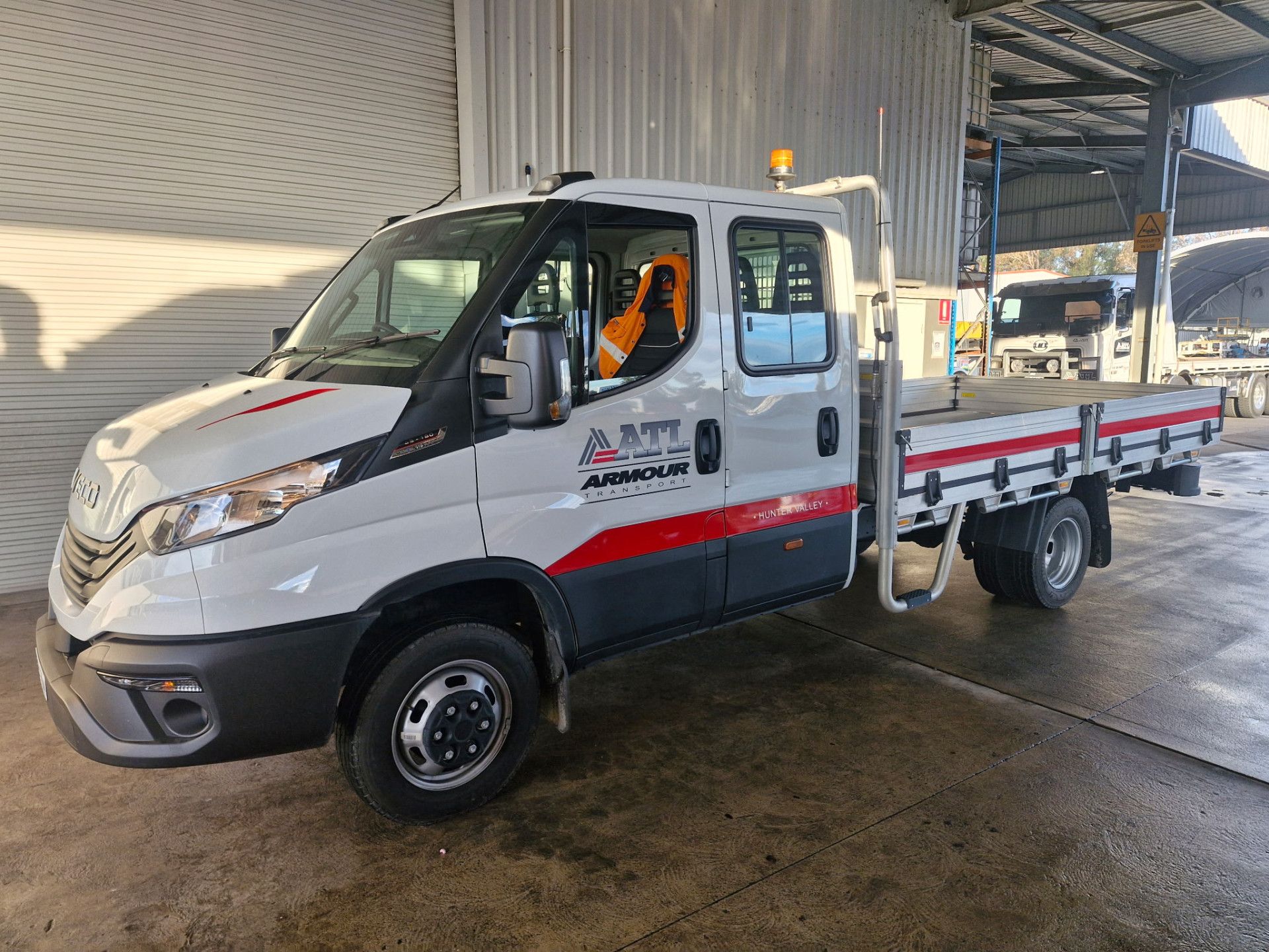 White flatbed utility truck parked inside a warehouse, with red and blue stripes and an amber roof light. -Armour Transport Logistics In Rutherford, NSW