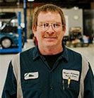 Mechanic in blue and gray uniform, glasses, smiling in a garage setting with a car lift. | Emge's Car Craft Auto Services