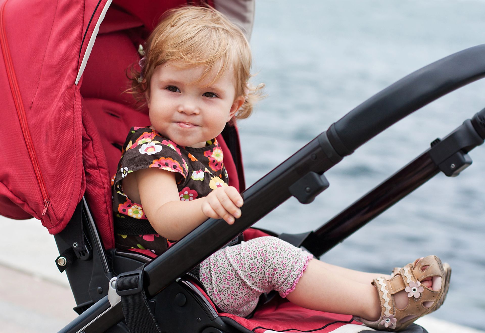 Bambina seduta in un passeggino rosso, con indosso una maglietta a fiori e leggings a fantasia, che guarda in camera.