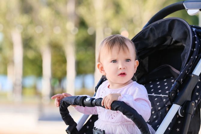 Bambino piccolo in un passeggino nero che si aggrappa alla sbarra, guarda avanti all'esterno con uno sfondo sfocato di un parco.