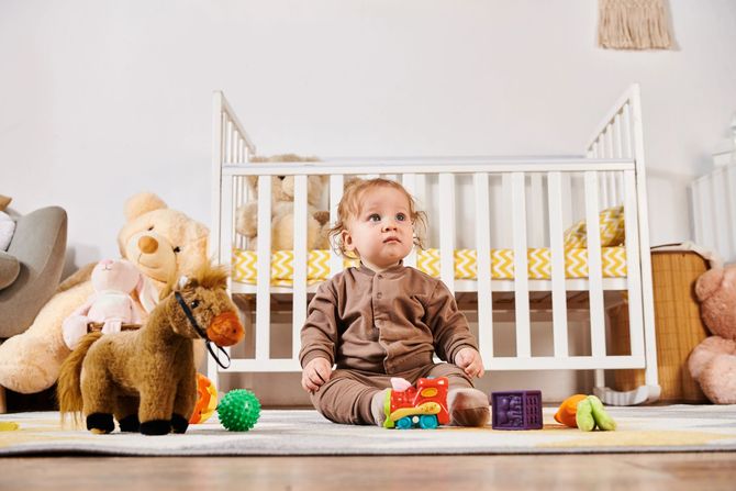 Un bambino piccolo è seduto sul pavimento accanto a una culla bianca, circondato da peluche e giocattoli colorati nella sua cameretta.