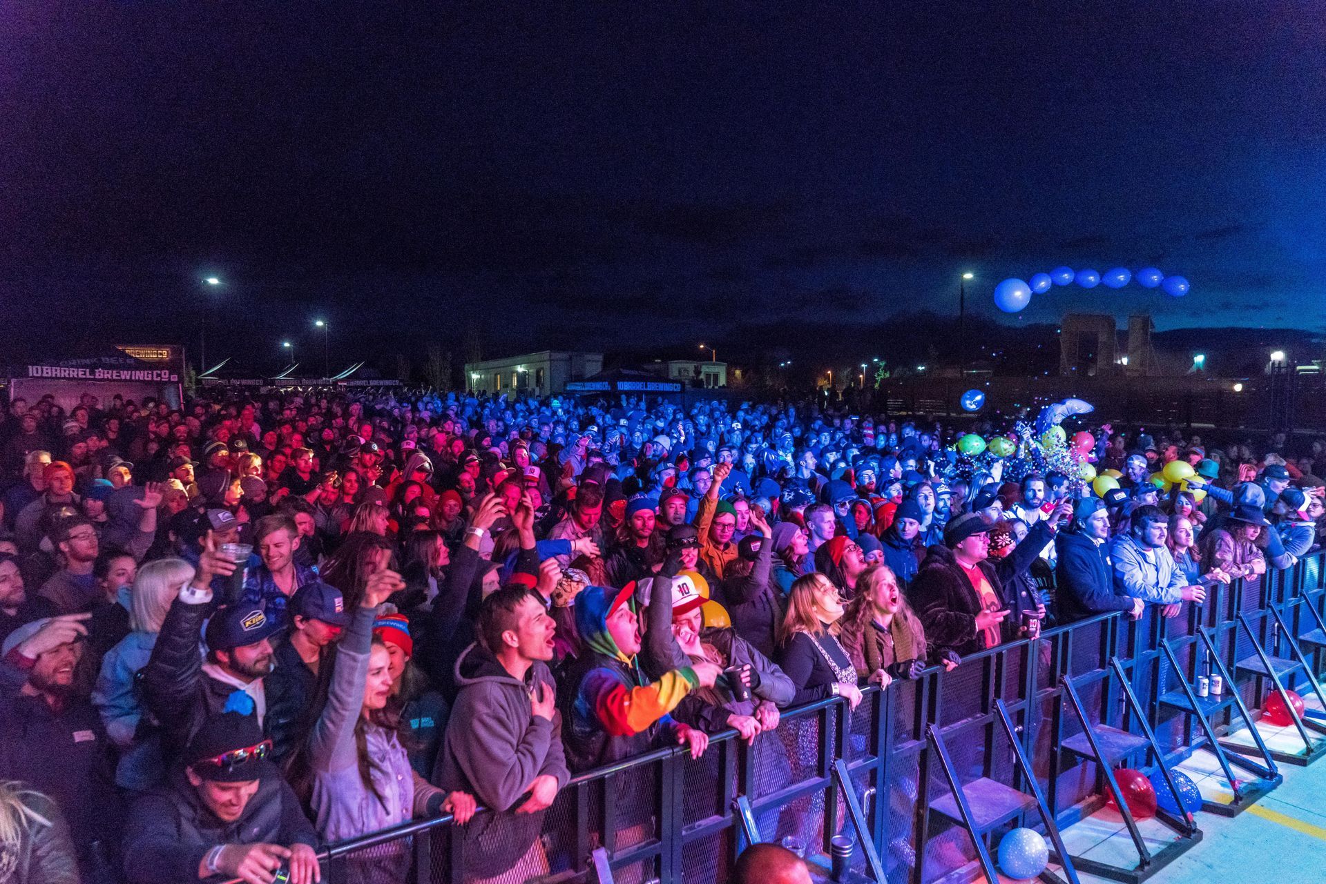 Crowd at an outdoor concert behind barriers, lit by colorful blue and red stage lights at night