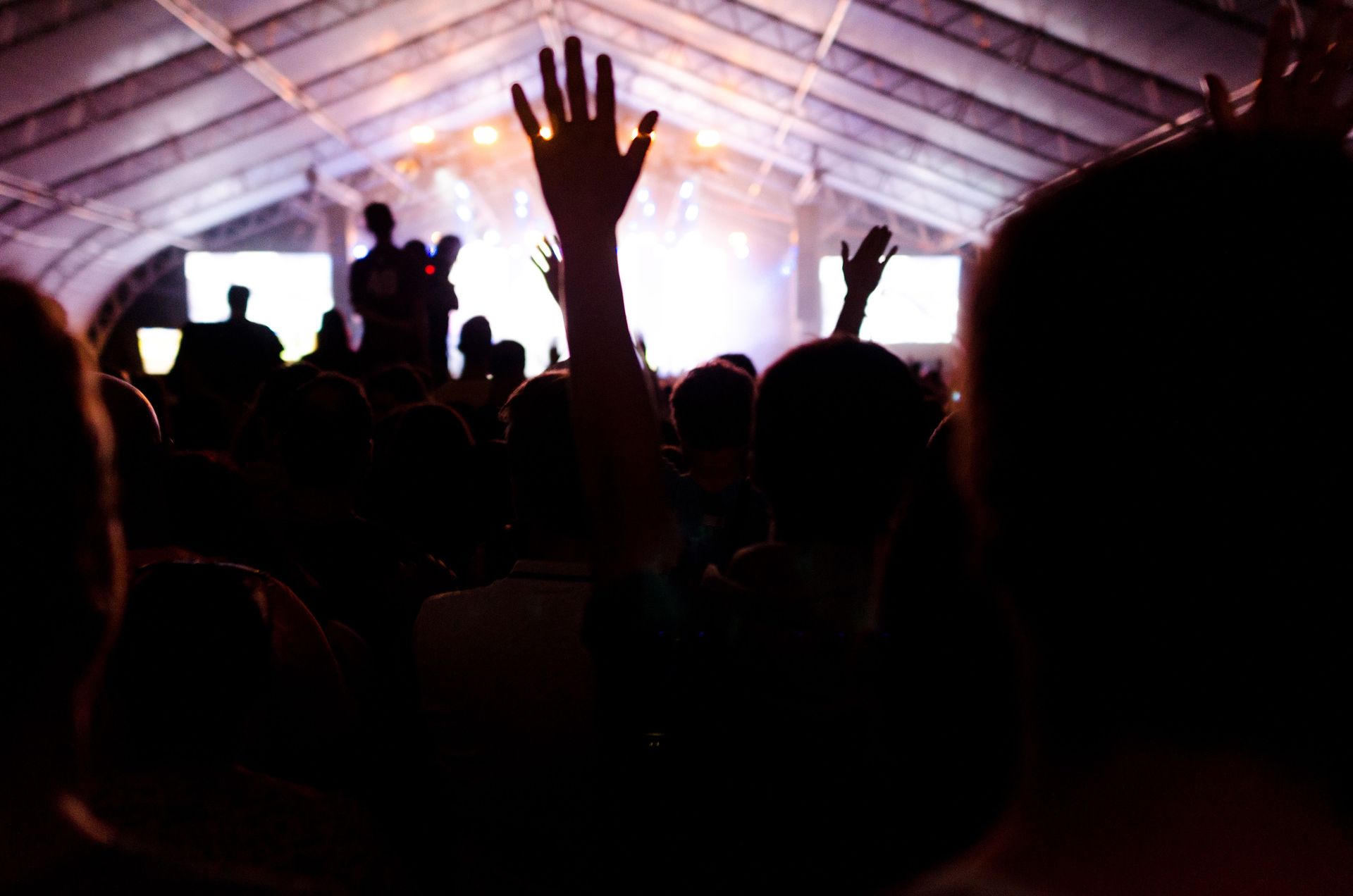 Crowd silhouettes with raised hands at a bright concert inside a tent