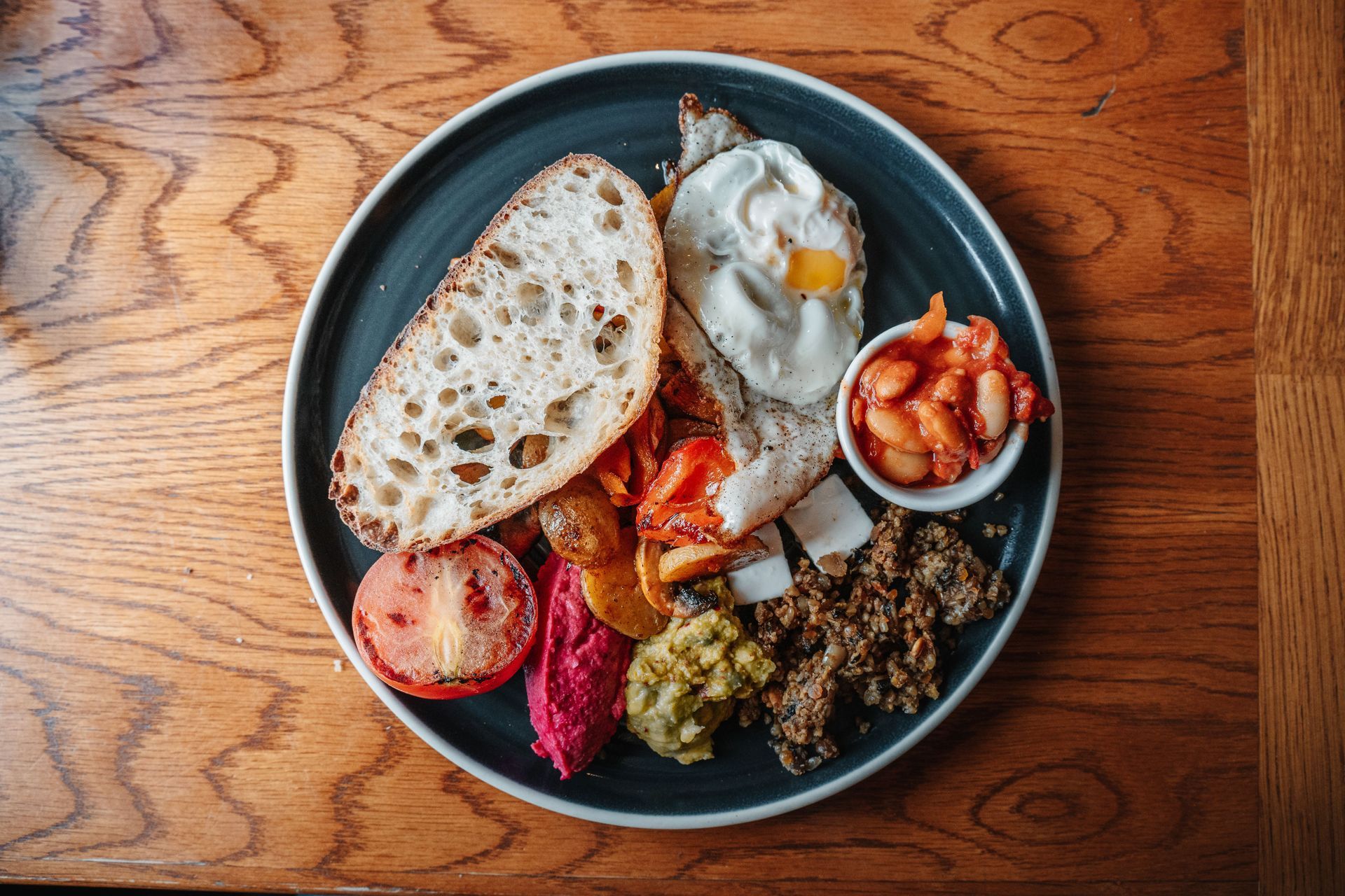 Plate of assorted brunch foods, including bread, eggs, beans, and vegetables, on a wooden table